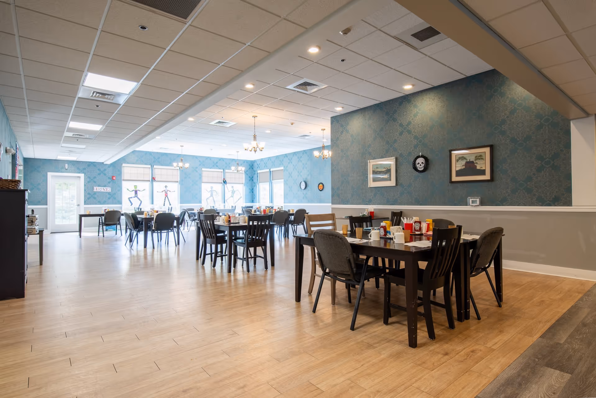 A spacious dining room with multiple tables and chairs arranged neatly. The tables are set with condiments, cups, and utensils. The room has light wood flooring, blue patterned wallpaper, and several framed pictures on the walls. Large windows allow natural light to brighten the space, and ceiling lights provide additional illumination.