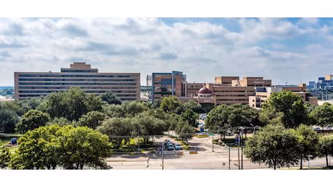 Wide exterior view of a large multi-story building complex surrounded by many trees and greenery under a partly cloudy sky.