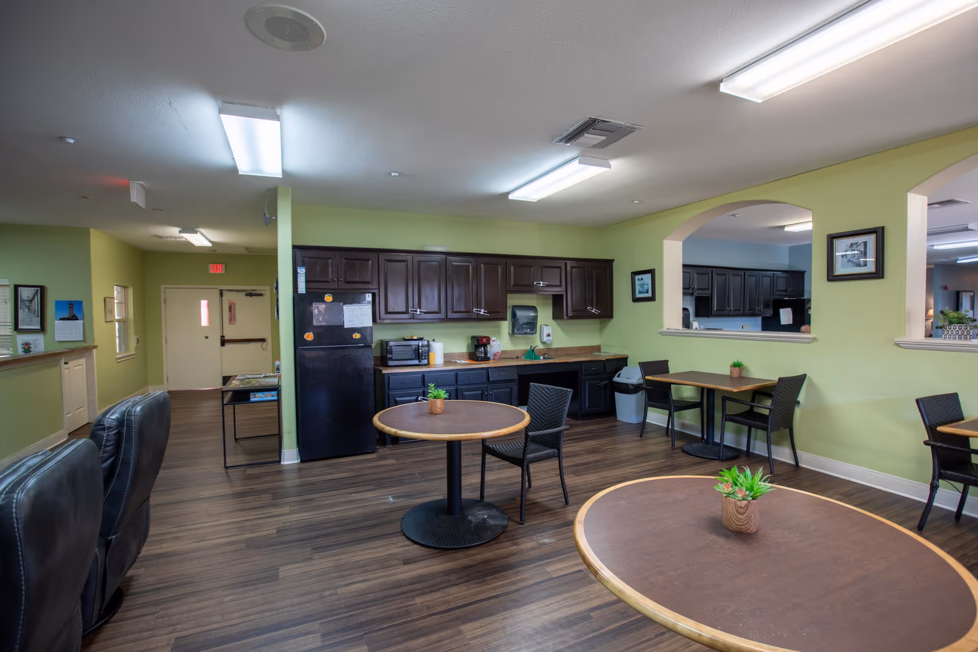 Communal dining area with round tables and chairs in front of a kitchenette with dark cabinets and green walls.