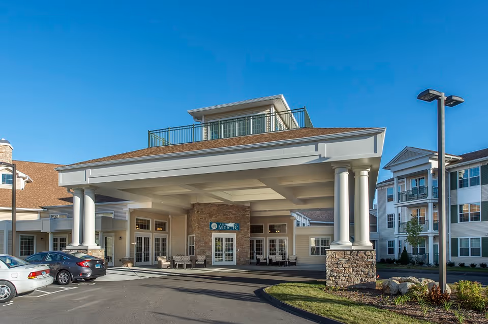 Front exterior of Masonicare At Mystic showing a covered entrance with large columns, parked cars and a clear blue sky.