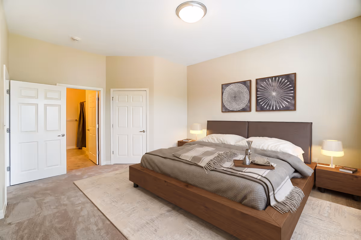 A modern bedroom with a large wooden bed featuring gray bedding and pillows. Two framed abstract artworks hang above the headboard. There are two bedside tables with lamps on each side of the bed. The room has beige walls and carpeted flooring with a light-colored area rug. Two white doors are visible, one leading to a bathroom with a towel hanging inside.