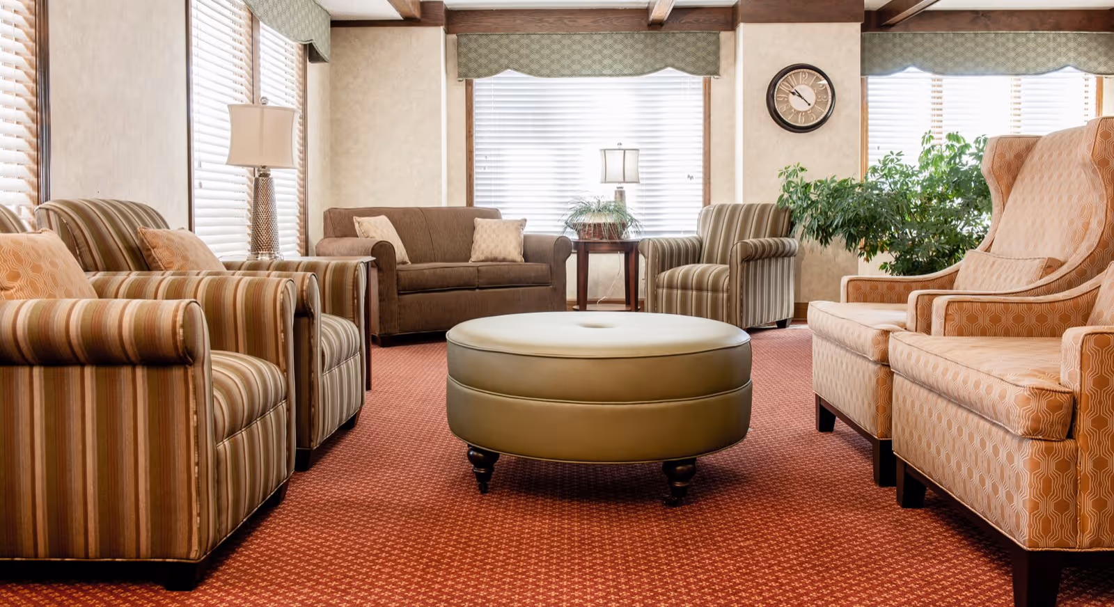 Comfortable senior living common room with upholstered armchairs, sofas, a round ottoman, lamps and large windows.
