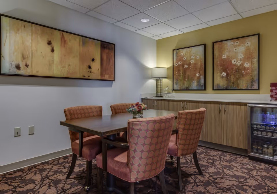 A cozy dining area with a rectangular wooden table surrounded by four upholstered chairs with a circular pattern. The room features a patterned carpet, a sideboard with cabinets, a small lamp, and two framed floral paintings on the wall. A small refrigerator with beverages is visible on the right side.