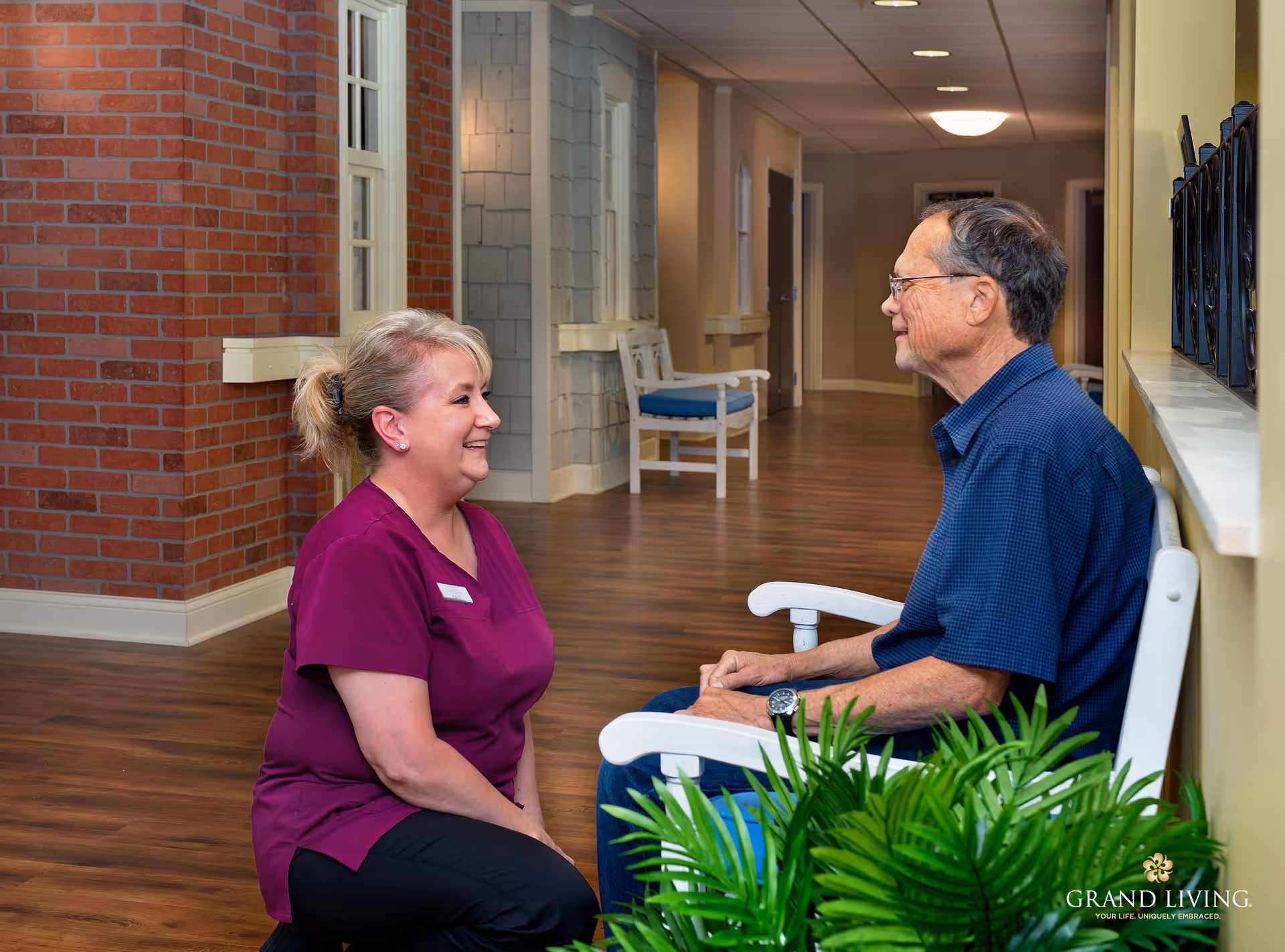 A female caregiver in a purple uniform kneels and smiles while talking to an elderly man sitting on a white bench in a hallway with wooden floors, brick and beige walls, and potted plants.
