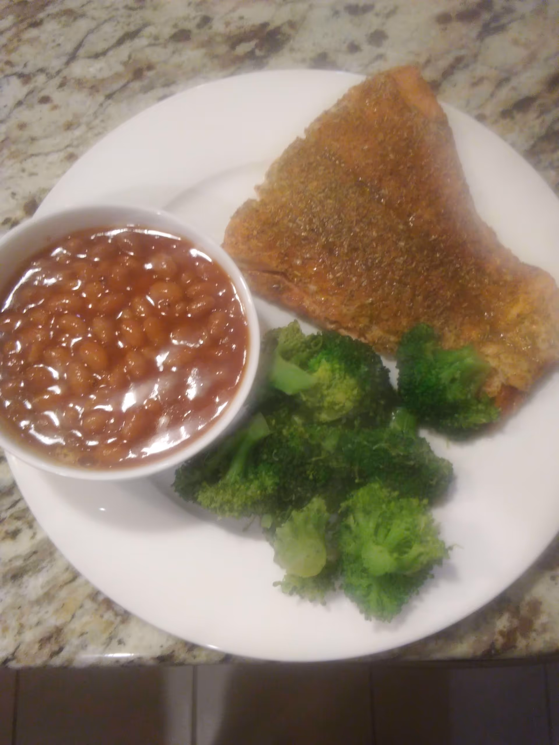 A white plate with a serving of baked beans in a small bowl, steamed broccoli florets, and a seasoned piece of cooked fish, placed on a granite countertop.