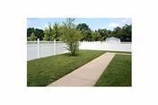 A paved walkway runs through a grassy yard bordered by a white vinyl fence with a small tree near the center. Trees and shrubs are visible beyond the fence under a partly cloudy sky.