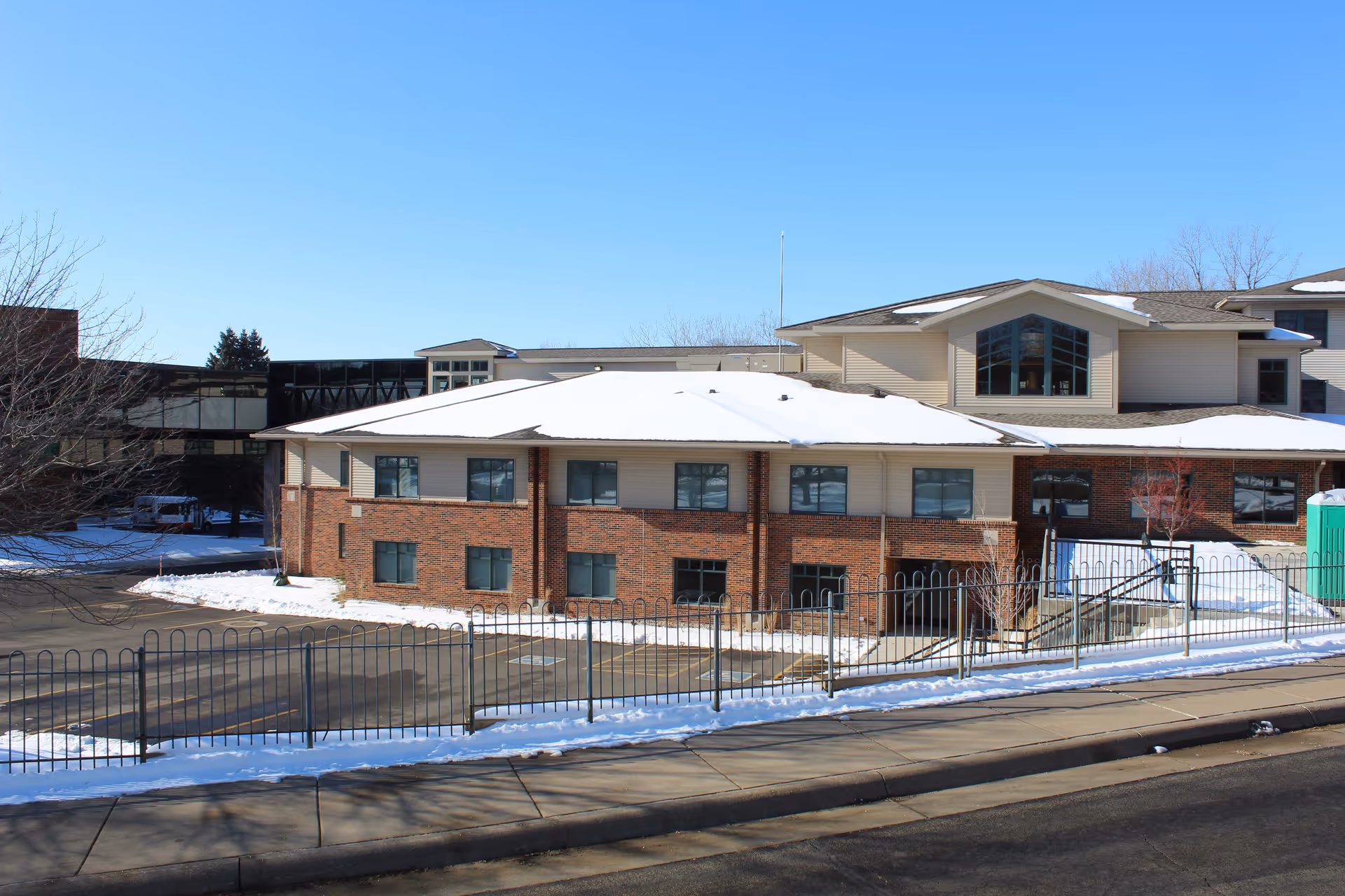 Exterior front view of a multi-story brick-and-siding senior living building with a snow-covered roof and a fenced driveway.