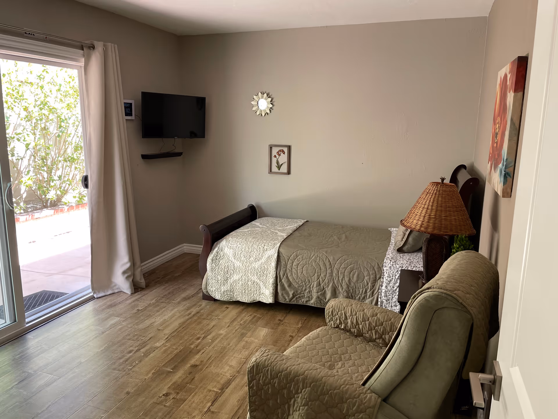 A senior living bedroom with a single bed covered in a green quilt and a white blanket, a green armchair with a quilted cover, a wooden nightstand with a wicker lamp, a wall-mounted flat screen TV, and a sliding glass door with curtains leading to an outdoor area with greenery.