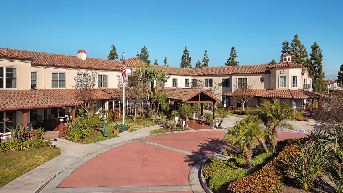 Exterior view of a senior living facility with a two-story building featuring a red tile roof, beige walls, and multiple windows. The entrance has a covered porch with benches and is surrounded by landscaped greenery, palm trees, and a circular driveway. An American flag is displayed near the entrance under a clear blue sky.