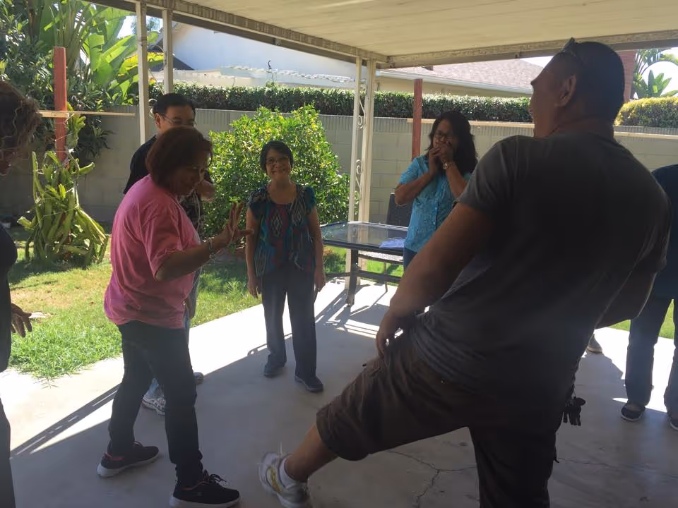 A group of people standing and interacting under a covered patio area with a table in the background. The setting is outdoors with greenery and a concrete floor visible.