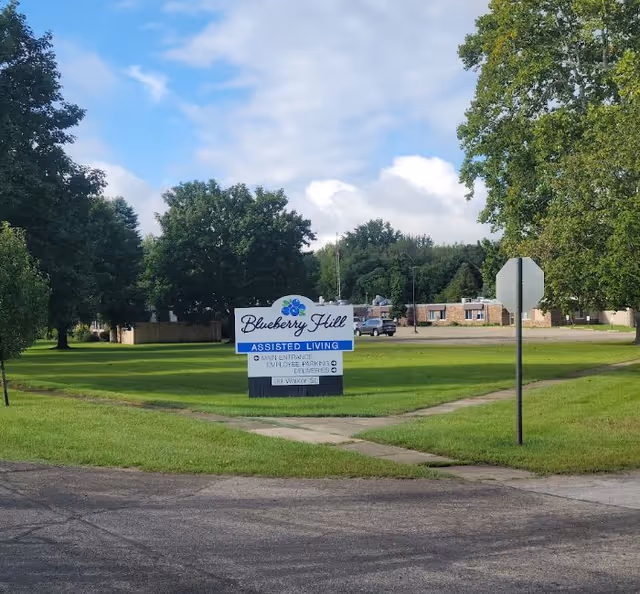 Front lawn with a sign reading "Blueberry Hill Assisted Living" and the facility building and trees in the background.