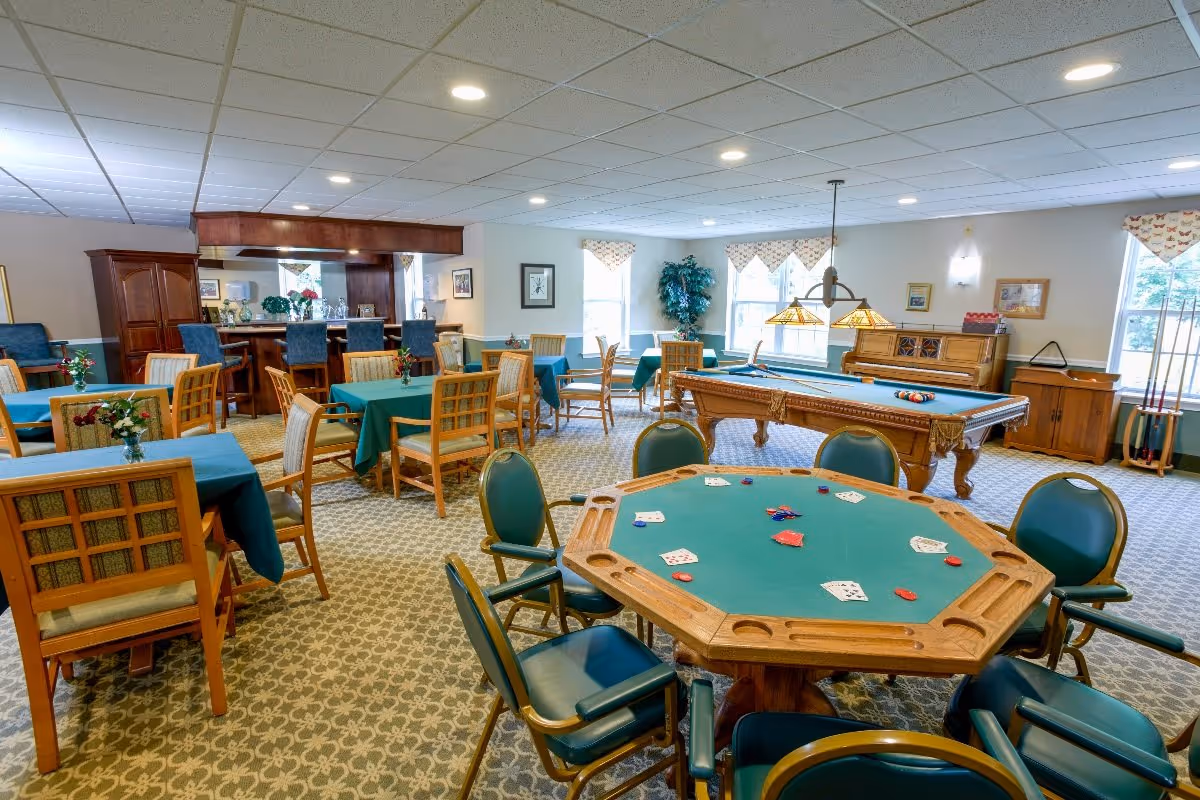 A spacious activity room in a senior living facility featuring a poker table with playing cards and chips, a pool table with billiard balls, multiple tables with green tablecloths and wooden chairs, a bar area with high chairs, and large windows letting in natural light.