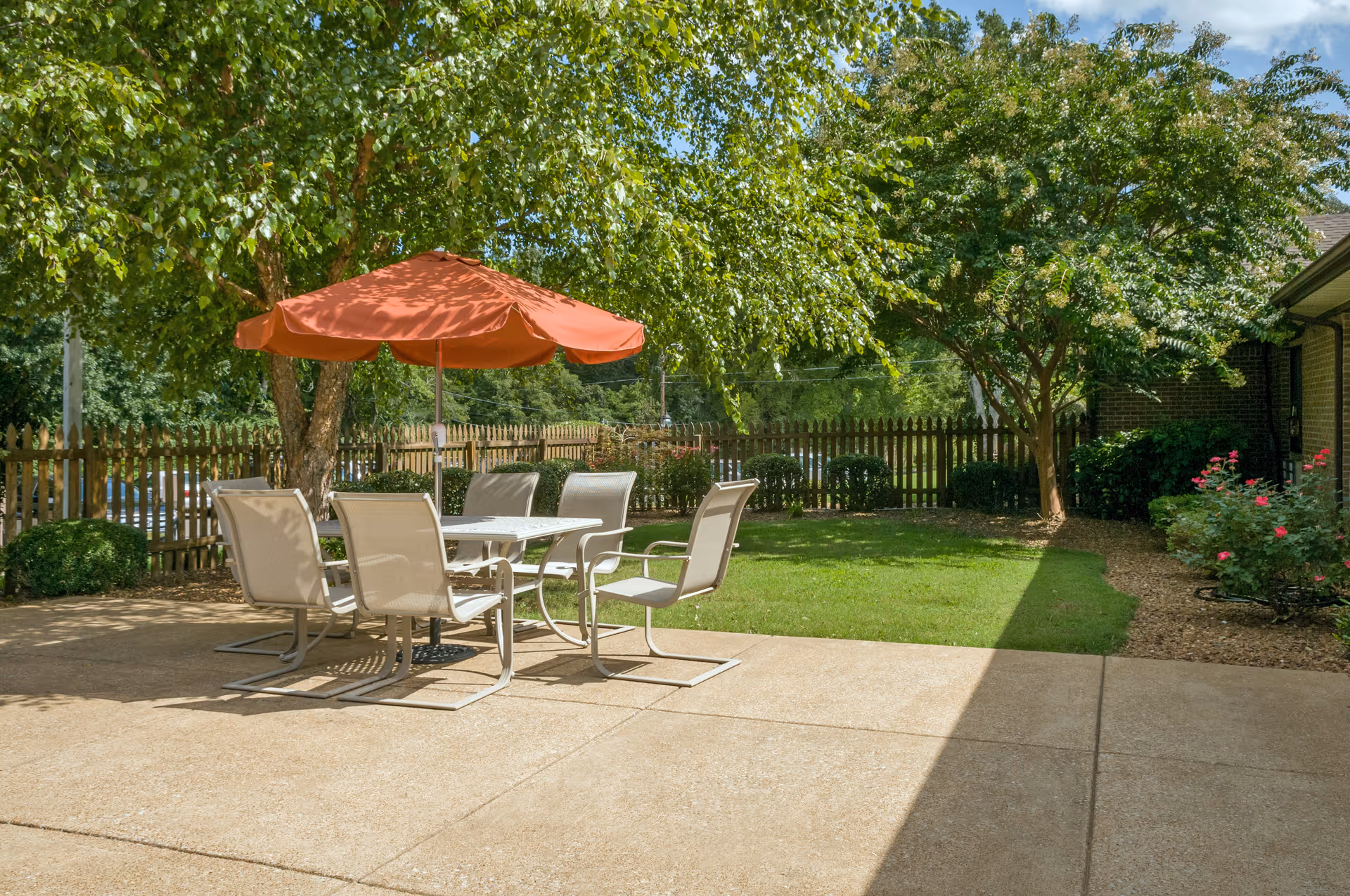 Patio with a table, six chairs and a red umbrella overlooking a grassy yard with trees and a picket fence.