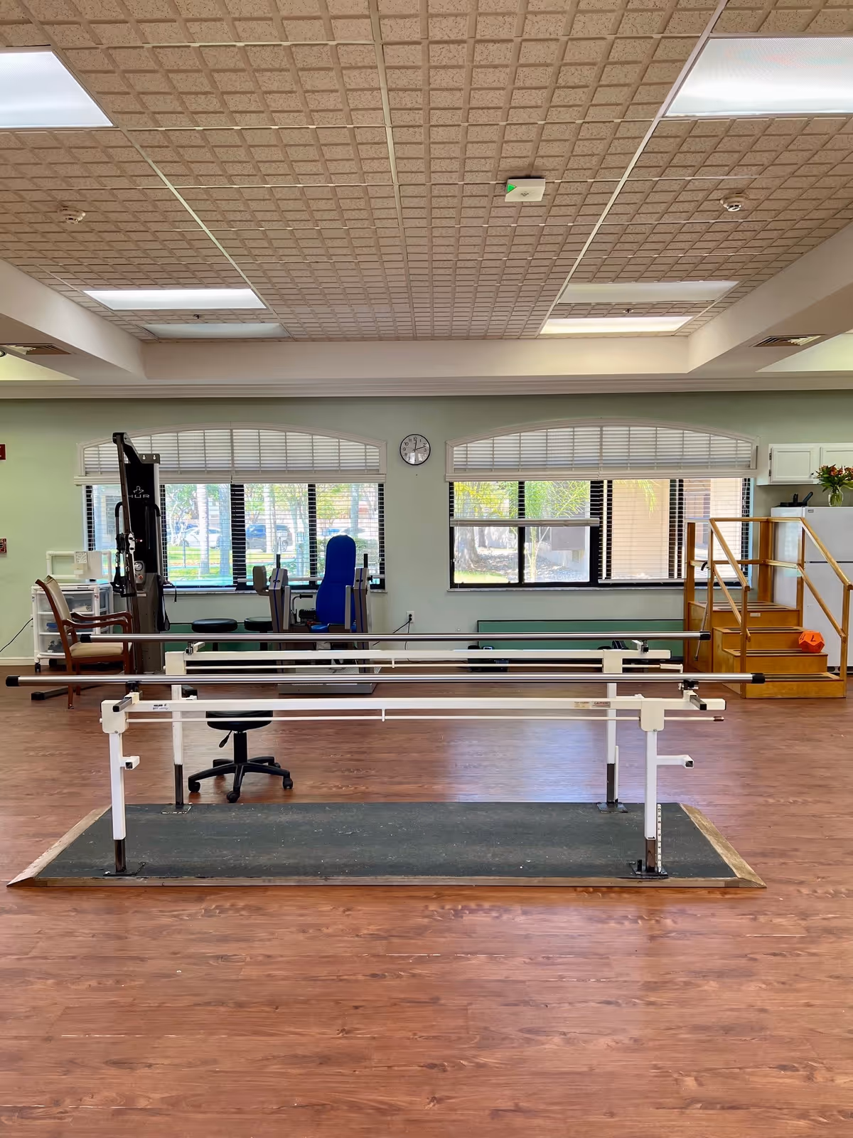 A rehabilitation gym room with parallel walking bars in the center, exercise equipment, windows and a small set of wooden stairs.