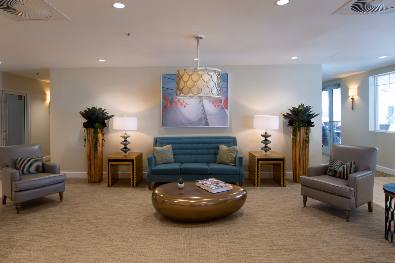 Communal lounge area with a teal sofa, two gray armchairs, side tables with lamps, decorative plants and a low coffee table.