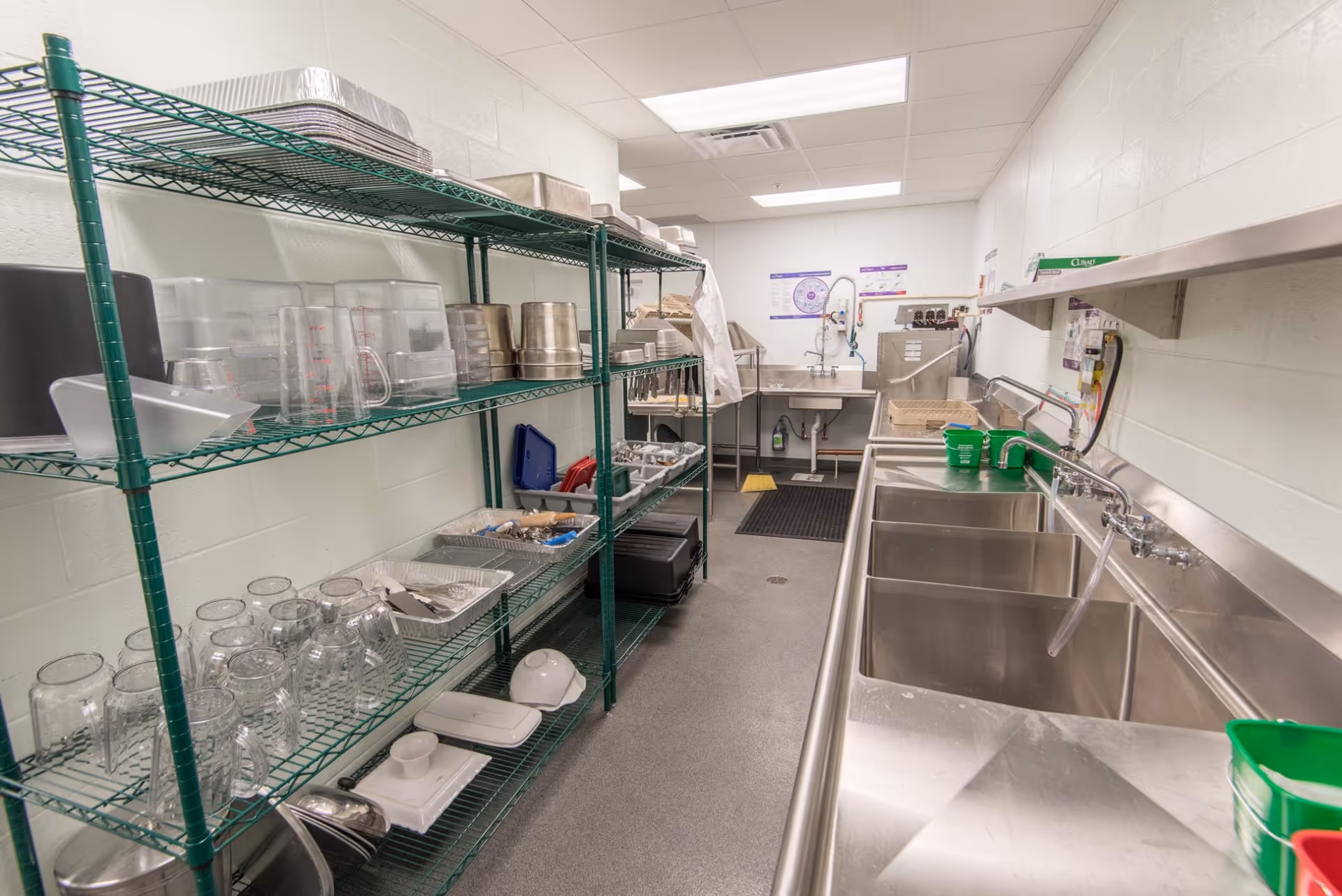 A clean and organized commercial kitchen dishwashing area with stainless steel sinks on the right and green metal shelving on the left holding various kitchen utensils, glass mugs, trays, and containers. The room has white walls and a gray floor with fluorescent lighting overhead.