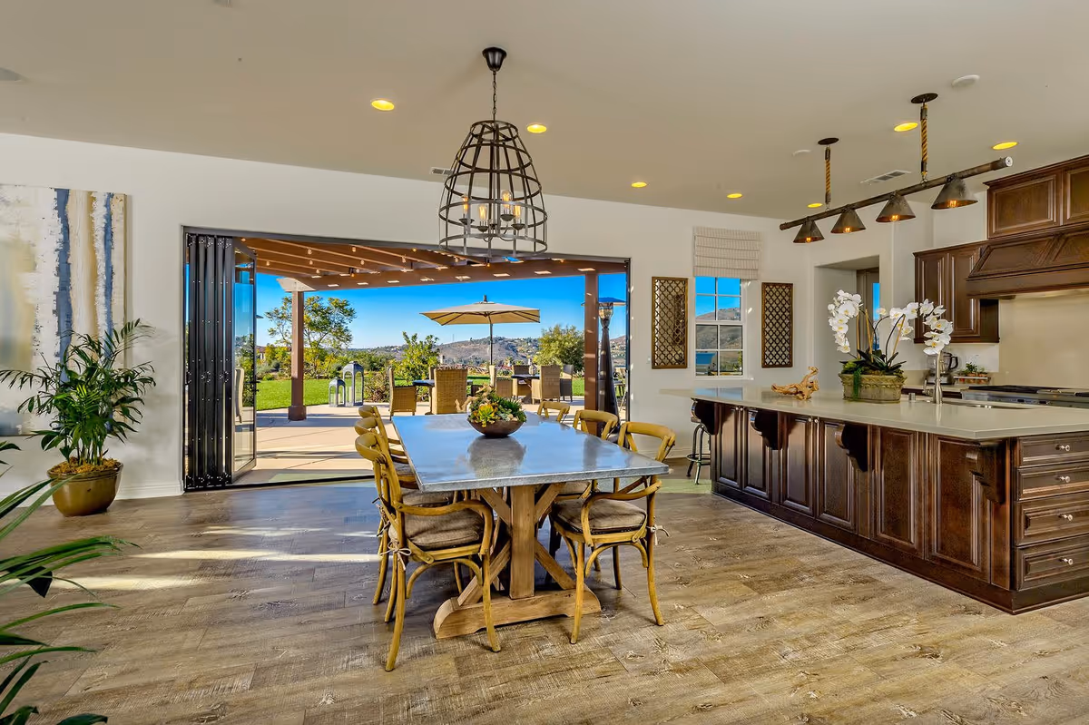 Bright and spacious open-plan kitchen and dining area with a large wooden dining table surrounded by six chairs. The kitchen features dark wood cabinetry, a large island with a white countertop, and pendant lighting. Large folding glass doors open to an outdoor patio with seating, an umbrella, and a scenic view of greenery and hills under a clear blue sky.