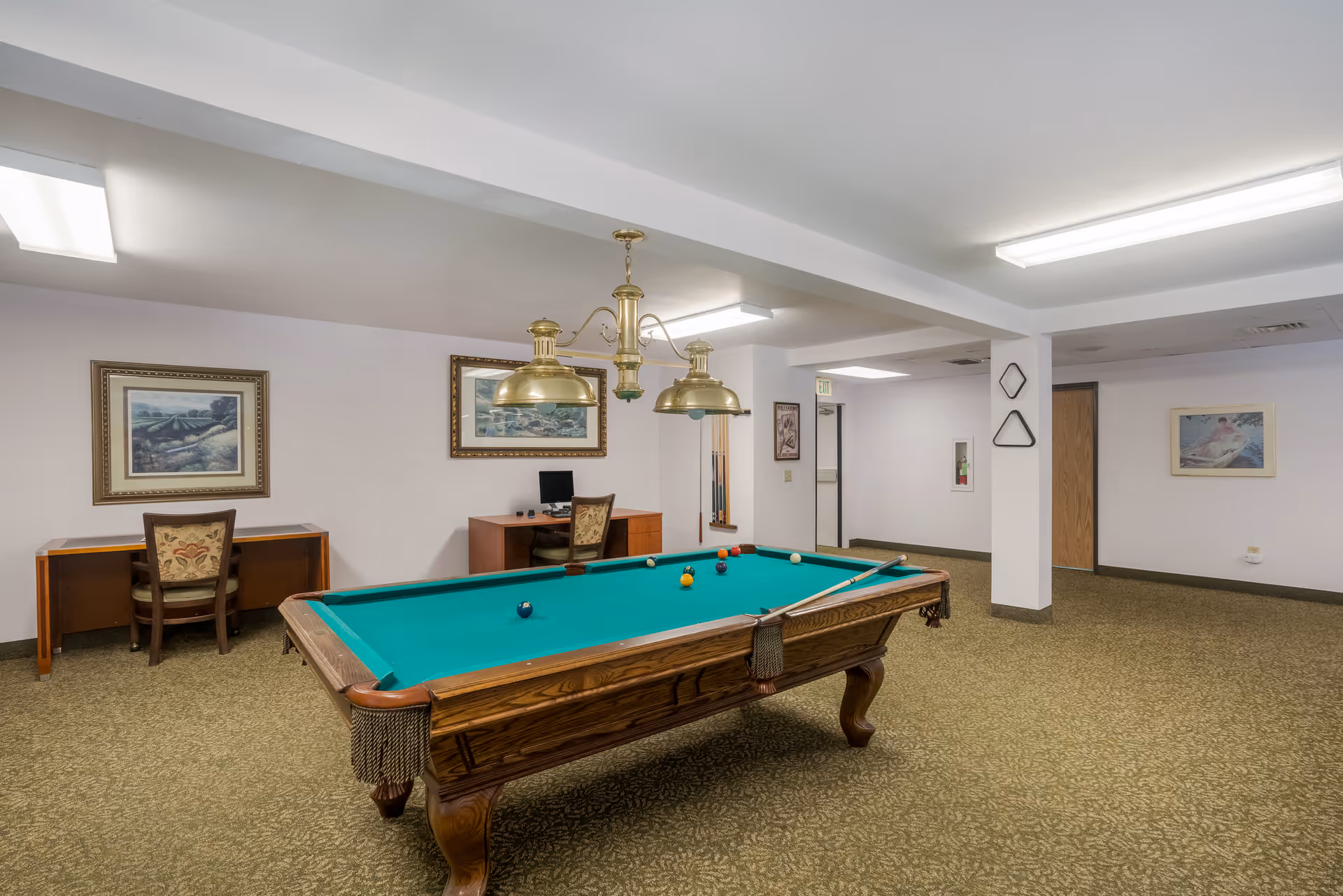A recreational room with a pool table in the center, a green felt surface with several billiard balls and cues on it. Above the table hang two brass pendant lights. Against the wall are two desks with chairs, one desk has a computer on it. The walls are decorated with framed paintings, and the floor is carpeted in a patterned design. The room has white walls and ceiling with fluorescent lighting.