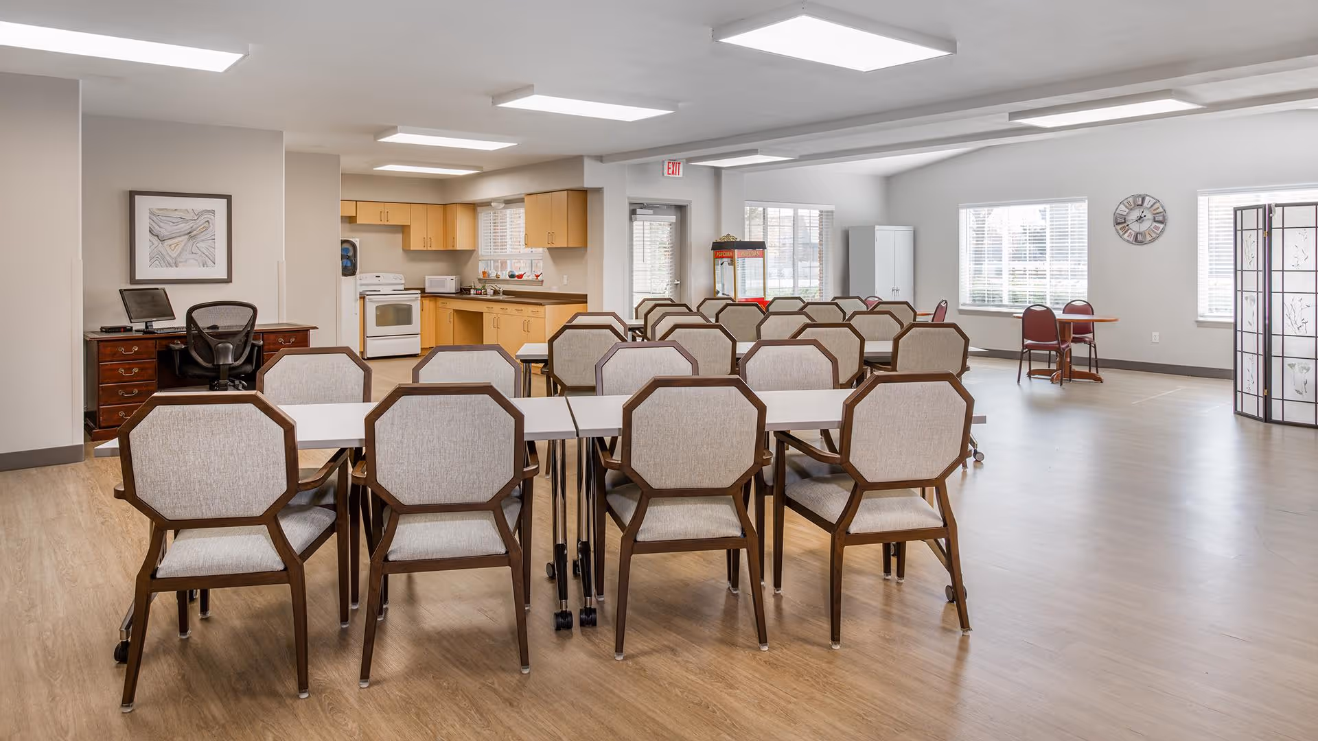 A spacious, well-lit common room with multiple tables and chairs arranged in rows. The room features a small kitchen area with light wood cabinets, a stove, microwave, and refrigerator. There is a desk with a computer and chair on the left side, large windows letting in natural light, a clock on the wall, and a decorative folding screen on the right side.