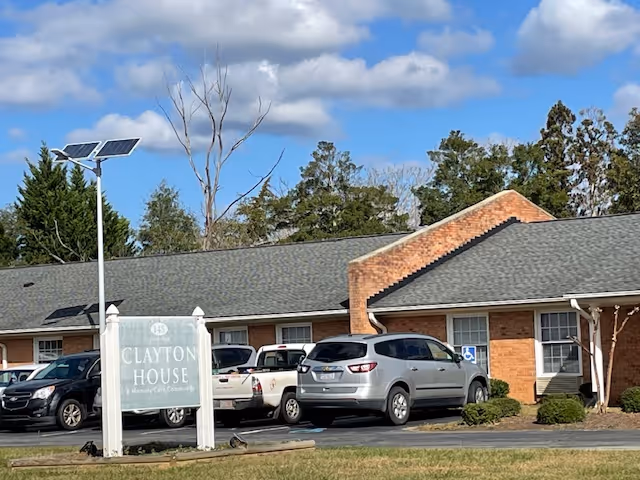 Exterior view of Clayton House, a single-story brick building with a gray shingled roof. Several cars are parked in front of the building, including a silver SUV and a white pickup truck. A sign in front reads 'Clayton House A Memory Care Community'. The sky is partly cloudy with some trees in the background.
