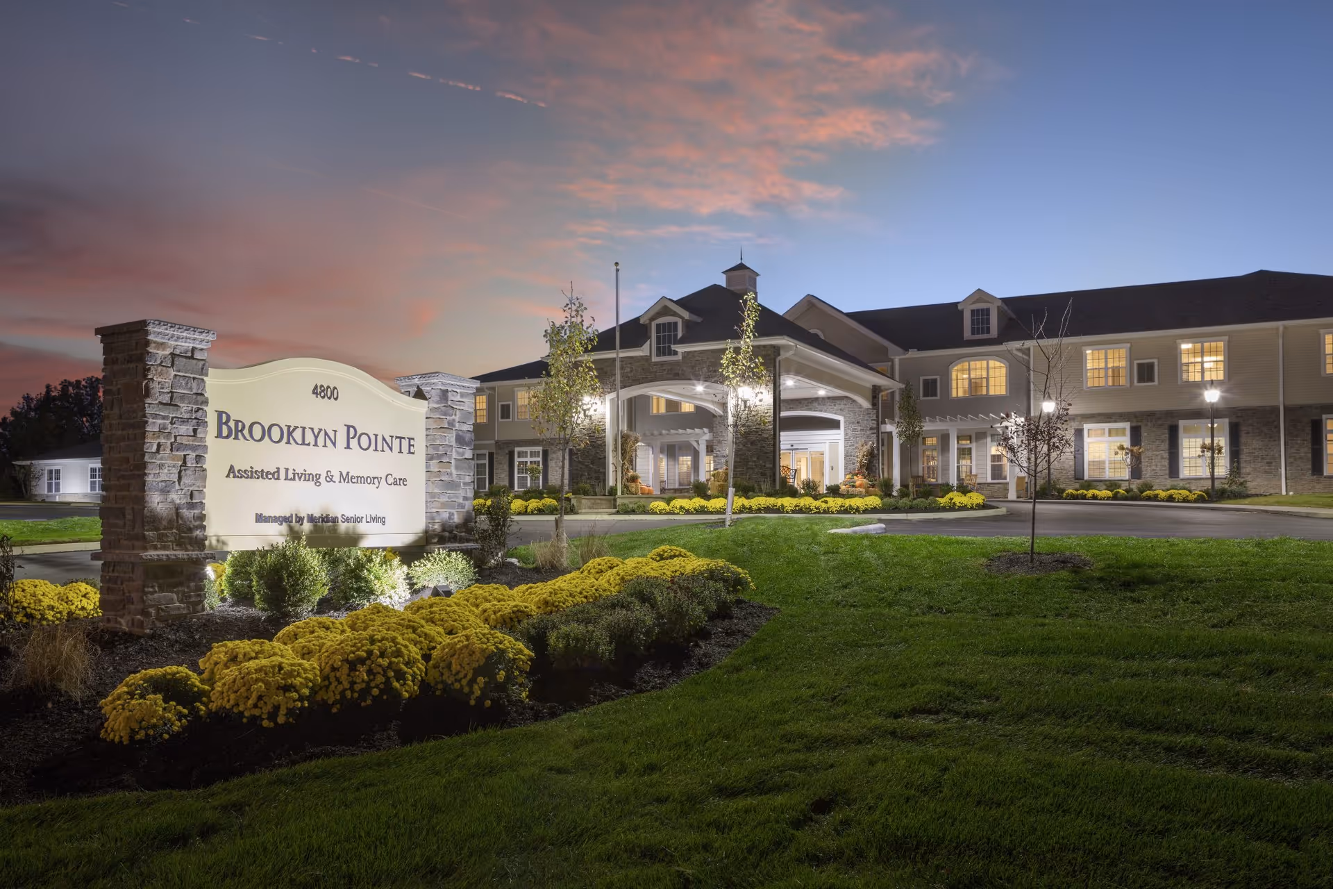 Exterior view of Brooklyn Pointe Assisted Living & Memory Care facility at dusk, showing the illuminated entrance, well-maintained landscaping with yellow flowers, and a large sign with the facility's name and address.