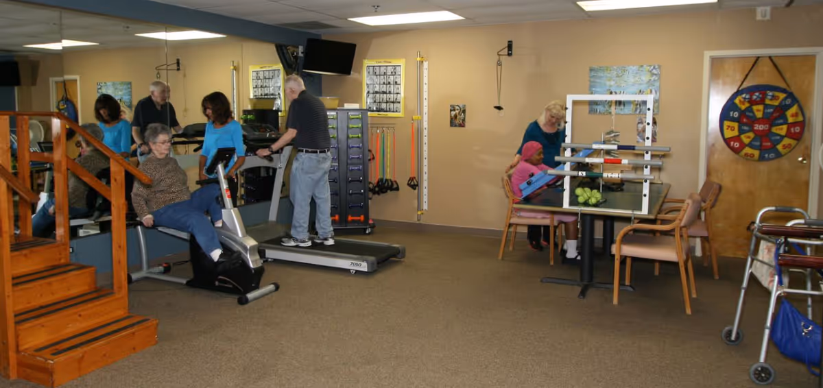 An indoor exercise room at a senior living facility with elderly individuals using fitness equipment. One person is on a recumbent bike, another is walking on a treadmill, and a caregiver is assisting a seated individual with arm exercises at a table. The room has beige walls, a large mirror, exercise bands, and a colorful target on a wooden door.