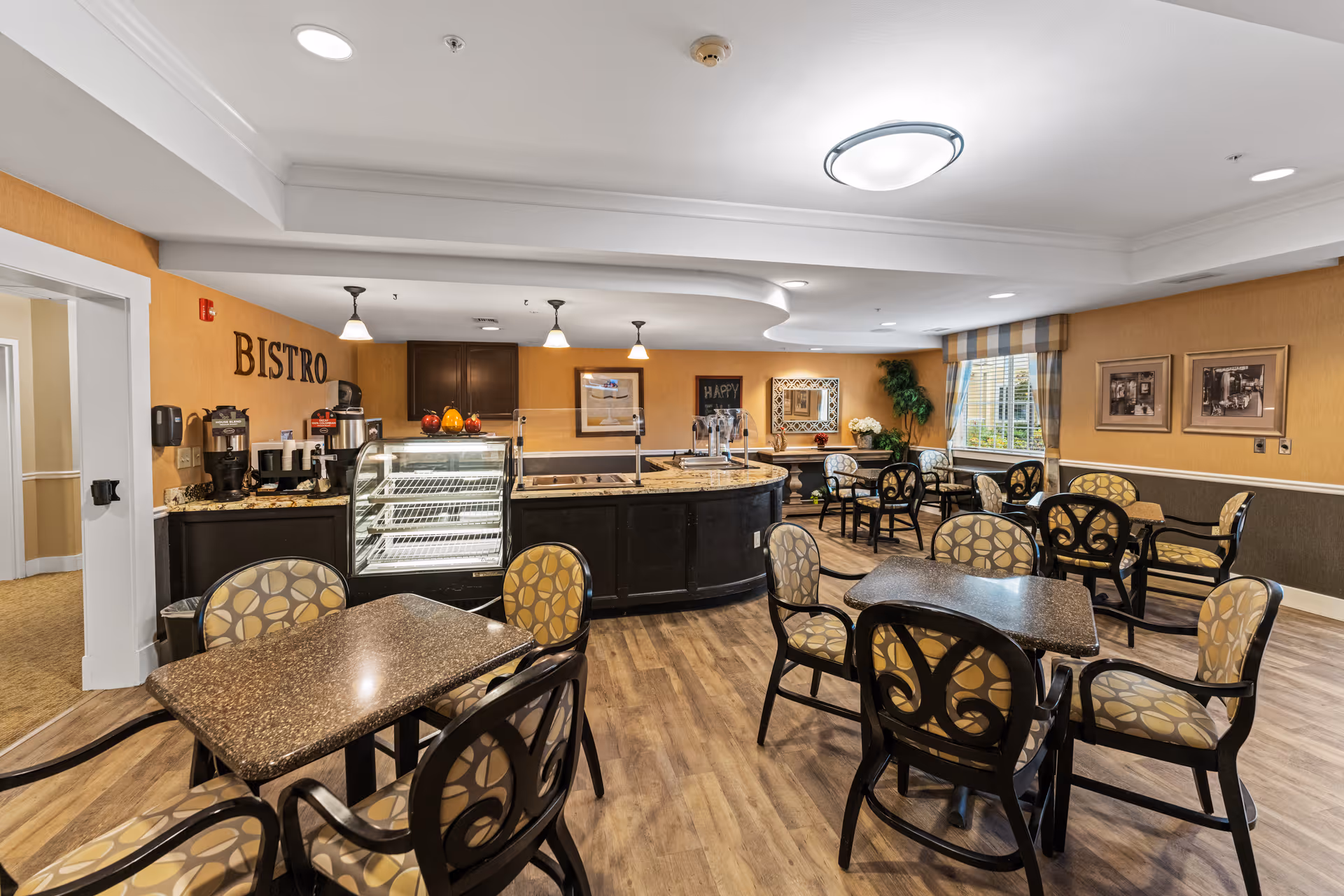 Interior view of a senior living facility bistro area with multiple tables and chairs arranged for dining. The room features warm yellow walls, wood flooring, and a counter with a glass display case and coffee machines. There are framed pictures on the walls, a window with striped curtains, and decorative plants.