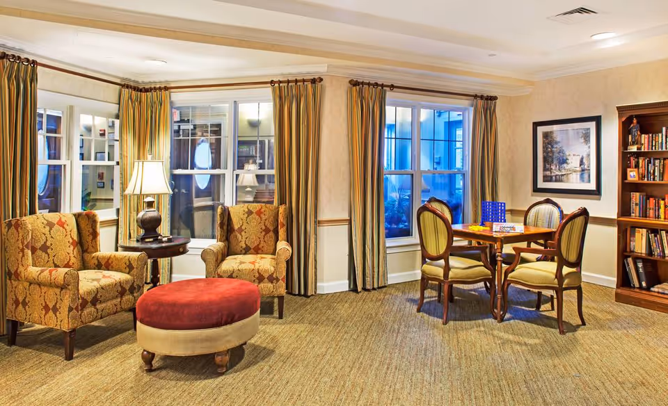 A cozy senior living common area with two patterned armchairs and a round ottoman near a small table with a lamp. Across the room, there is a wooden table with four chairs, a board game set up on the table, a bookshelf filled with books, and a framed picture on the wall. The room has large windows with striped curtains and beige walls.