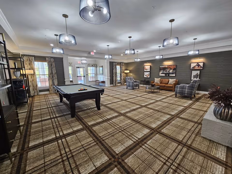 A spacious common area in an assisted living facility featuring a pool table in the center, a seating area with a brown leather sofa and two plaid armchairs against a dark accent wall with framed artwork, large windows with patterned curtains, multiple ceiling lights with modern fixtures, and a patterned carpet floor.