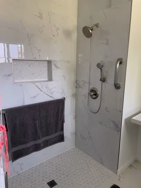 A modern shower area with white and gray marble-patterned tiles on the walls and small hexagonal tiles on the floor. The shower features a wall-mounted showerhead, a handheld showerhead, and a grab bar. There is a built-in recessed shelf in the wall and a dark gray towel hanging on a towel bar outside the shower area.