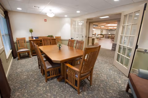 A well-lit dining room with a wooden rectangular table surrounded by eight wooden chairs with cushioned seats. The room has carpeted flooring with a patterned design, beige walls with white trim, and a sideboard against the far wall with a small plant and decorative items. French doors are open, leading to another room with additional seating and lighting fixtures.