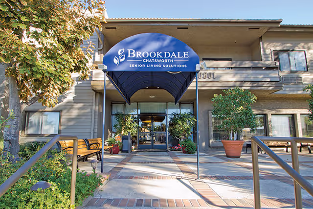 Entrance to Brookdale Chatsworth senior living facility with a blue canopy displaying the facility name and slogan. The entrance is flanked by potted plants and benches, with a tree and greenery on the left side.