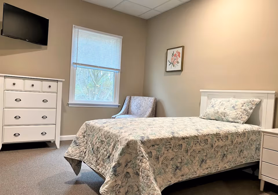 A bedroom with a bed covered in a floral quilt, a white dresser with drawers, a patterned armchair, a window with blinds, a wall-mounted TV, and a framed floral artwork on the wall.