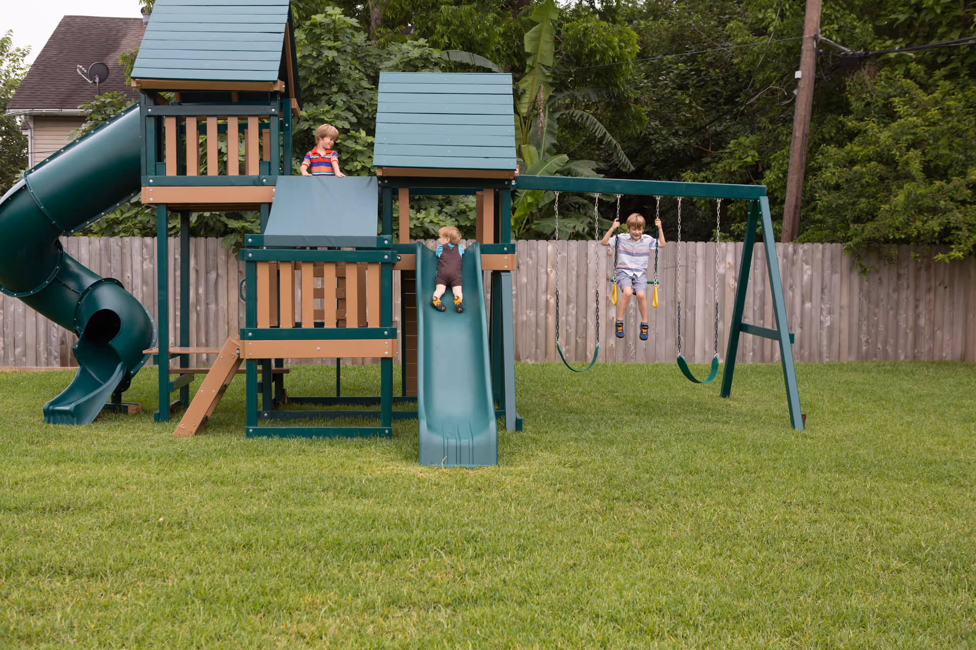 Three children playing on a green and brown playground set in a grassy backyard. One child is at the top of a slide, another is sliding down, and the third is swinging on a swing. There is a wooden fence and trees in the background.