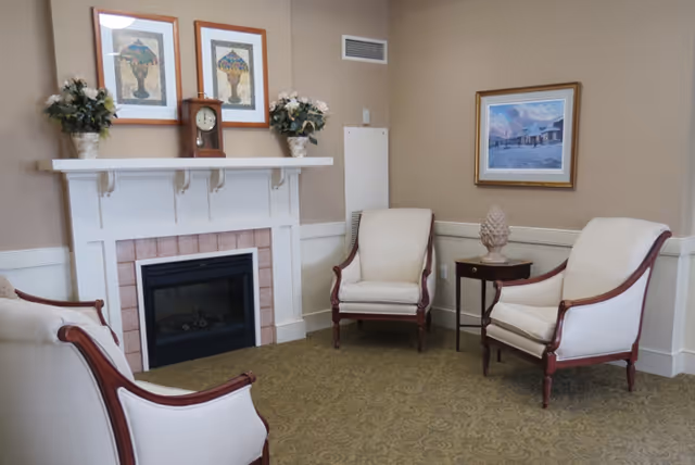 A cozy sitting area with four upholstered armchairs arranged around a white fireplace with a tiled surround. The mantel holds two framed pictures, a clock, and two flower arrangements. There is a small side table with a decorative item between two chairs, and a framed painting hangs on the beige wall.