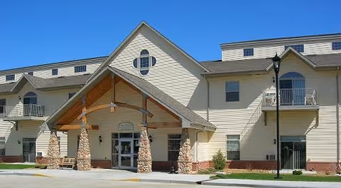 Exterior view of a multi-story senior living facility building with beige siding, stone pillars supporting a wooden entrance canopy, balconies, and a clear blue sky.