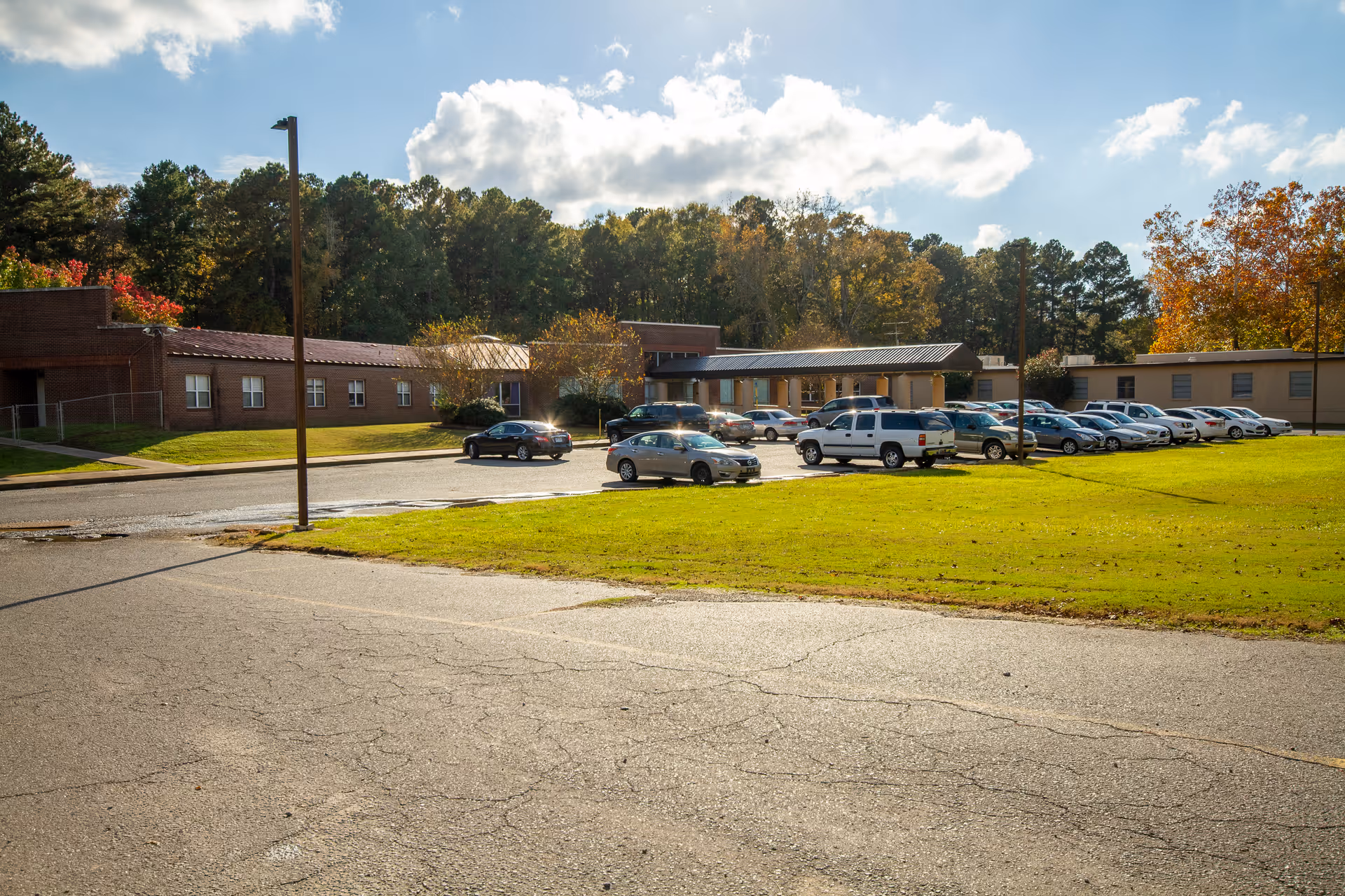 Exterior view of The Blossoms at West Dixon Assisted Living, Nursing Center & Rehab showing a parking lot with several cars parked, a grassy area, and a one-story building surrounded by trees under a partly cloudy sky.