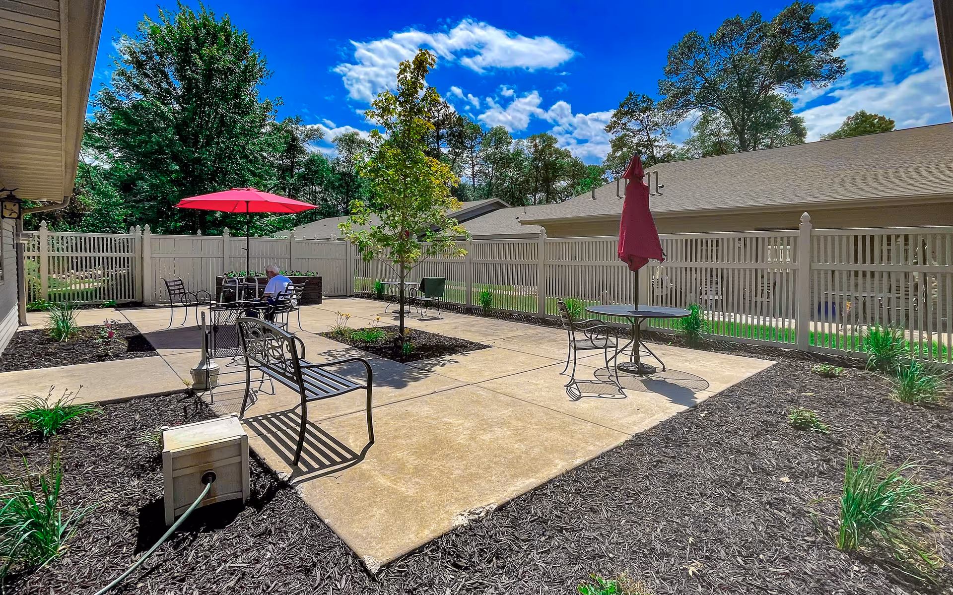 Outdoor patio area with concrete walkways, black metal benches, tables with red umbrellas, and a white fence surrounding the space. There are small landscaped garden beds with plants and a tree in the center. A person is sitting at one of the tables under an umbrella. The sky is blue with some clouds and trees are visible beyond the fence.
