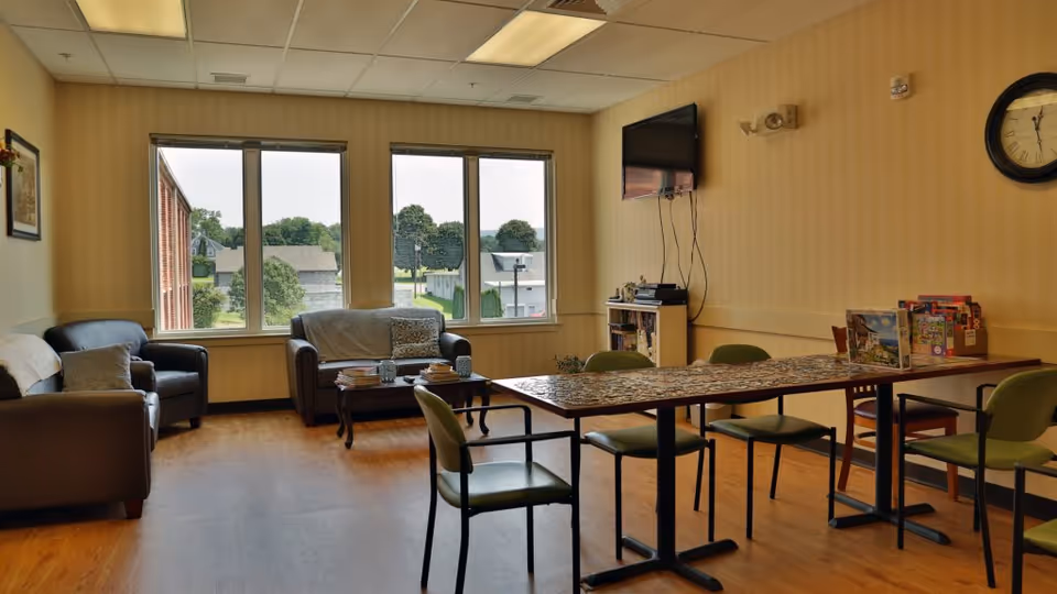 Communal living room with sofas by large windows, a long table with chairs and puzzles, and a wall-mounted TV.