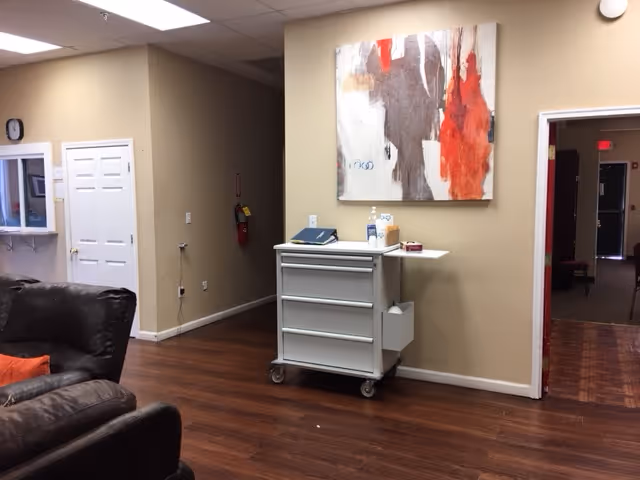 Interior view of a room with wooden flooring and beige walls. There is a brown leather couch with an orange pillow on the left side. A white rolling cart with drawers is positioned against the wall, holding various items including a bottle of hand sanitizer. Above the cart hangs an abstract painting with red, white, gray, and black colors. A doorway on the right leads to a dimly lit hallway, and a white door and a clock are visible on the left wall.