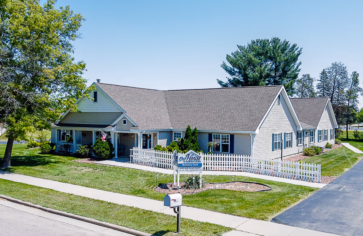 Single-story light-colored senior living memory care building with a white picket fence, front porch, and sign on a well-kept lawn.