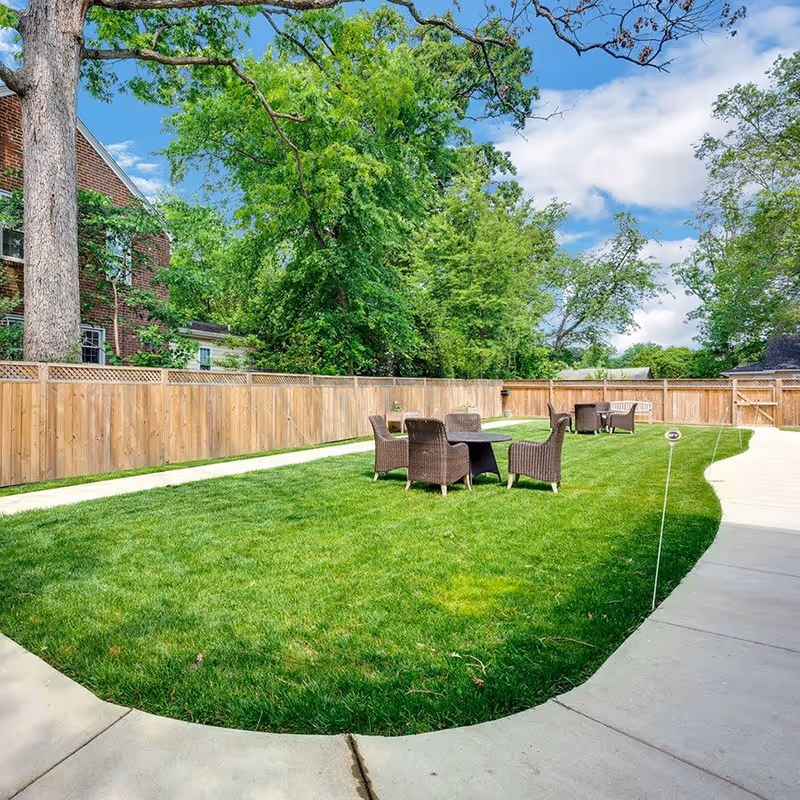 Outdoor area with green grass, a wooden fence, and several sets of brown wicker chairs and tables arranged on the lawn. There is a concrete pathway curving around the grass, and trees and a brick building are visible in the background under a partly cloudy blue sky.