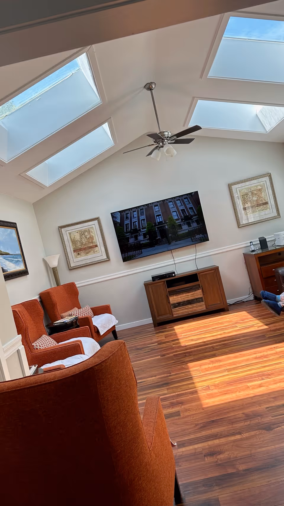 Sunlit living room with skylights, a ceiling fan, wall-mounted TV, and orange armchairs on wood flooring.