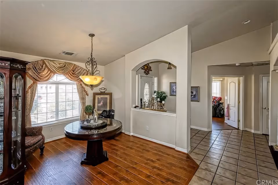 Interior view of a senior living facility showing a room with wooden flooring, a round wooden table with decorative items, a window with ornate curtains, and a hanging light fixture. Adjacent to this room is a tiled floor hallway leading to other rooms with open doors.