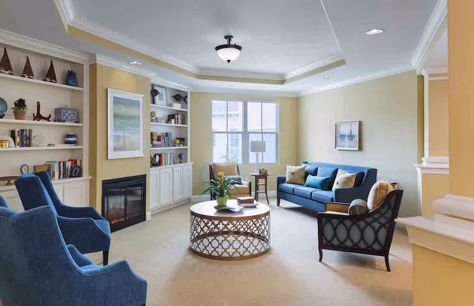 A bright and cozy living room with beige walls and carpet. The room features a blue sofa with multiple cushions, two blue armchairs, and a black armchair with a cushion. A round coffee table with a decorative metal base and a plant on top is centered in the room. Built-in white shelves with books and decorative items flank a fireplace on one wall. A window lets in natural light, and a ceiling light fixture is visible.