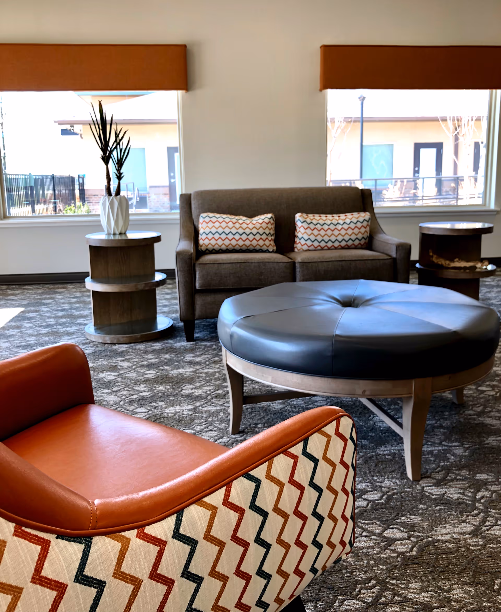 A cozy seating area in a living room with a brown sofa adorned with two patterned cushions, a round black leather ottoman, a patterned armchair with orange leather seating, and two wooden side tables with a white vase and plant. Large windows with orange valances allow natural light to fill the room.