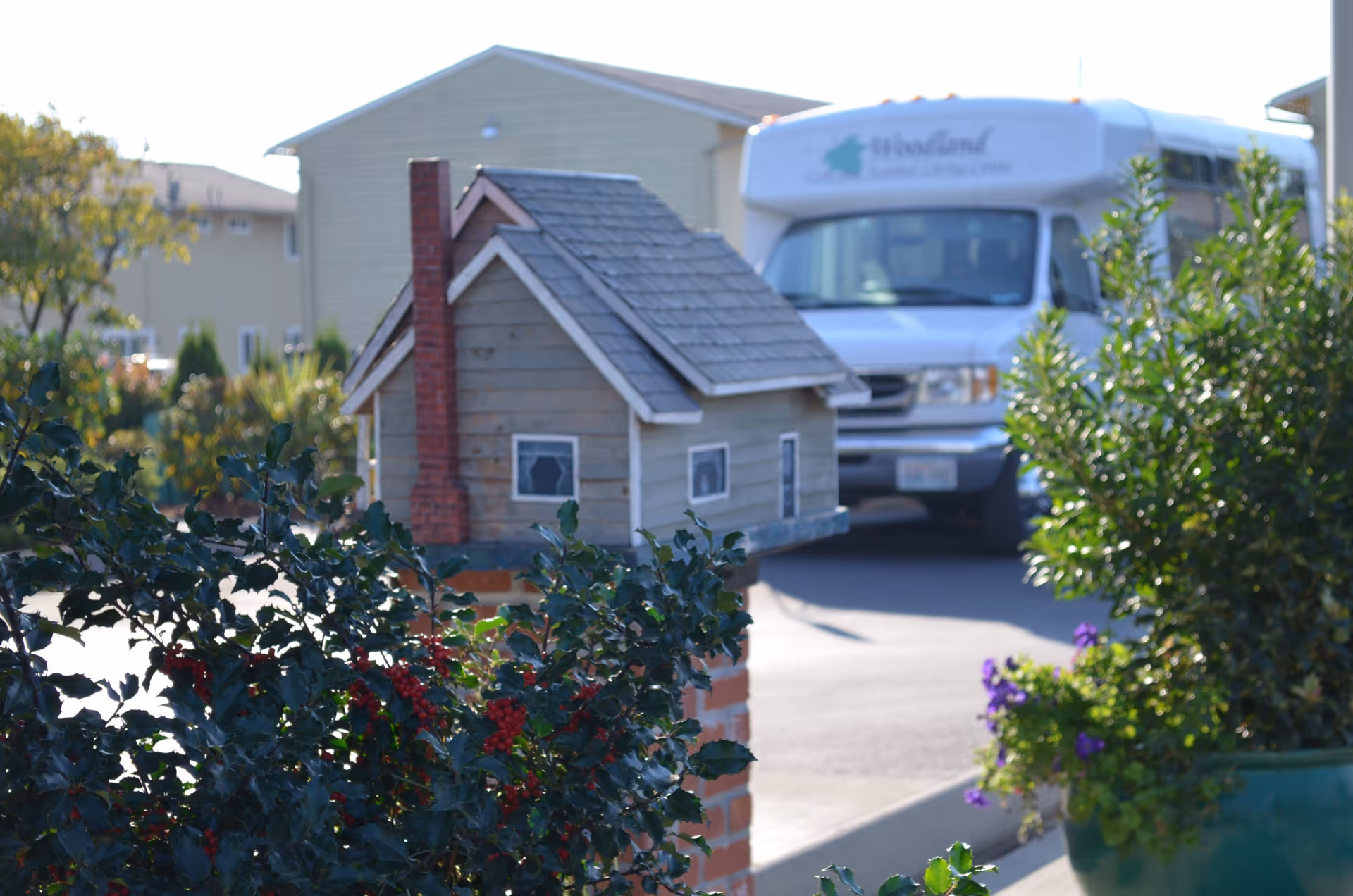 A small decorative wooden birdhouse with a chimney is mounted on a brick pillar surrounded by green bushes with red berries. In the background, there is a white vehicle with the Woodland Care Center & Assisted Living logo parked on a paved area.