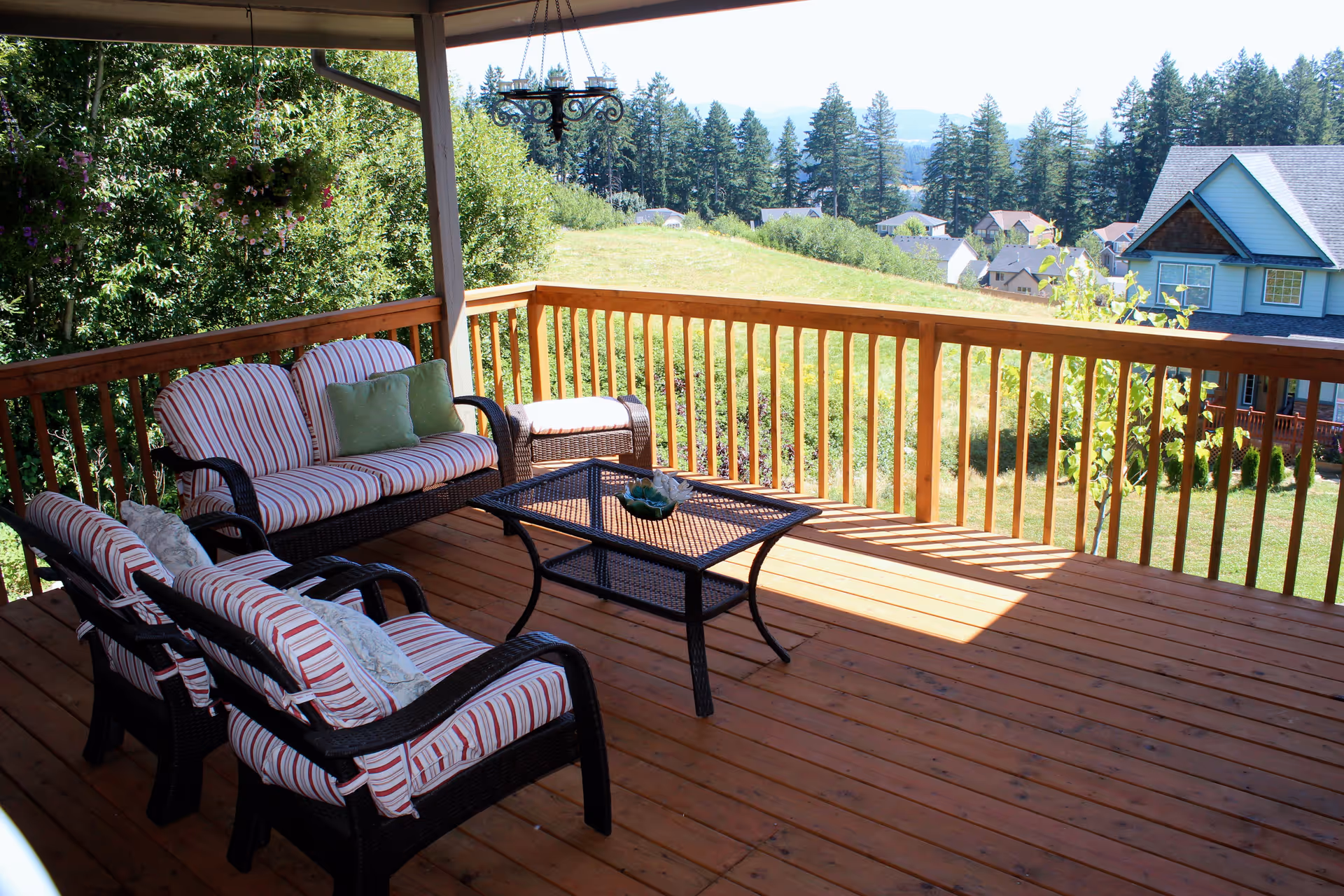 Covered wooden deck with outdoor wicker furniture including a striped cushioned loveseat, two chairs, an ottoman, and a glass-top coffee table. The deck overlooks a grassy hill, trees, and houses in the distance under a clear sky.