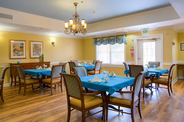 Dining room with several tables set with teal tablecloths and wooden chairs under a chandelier near windows and an exit door.
