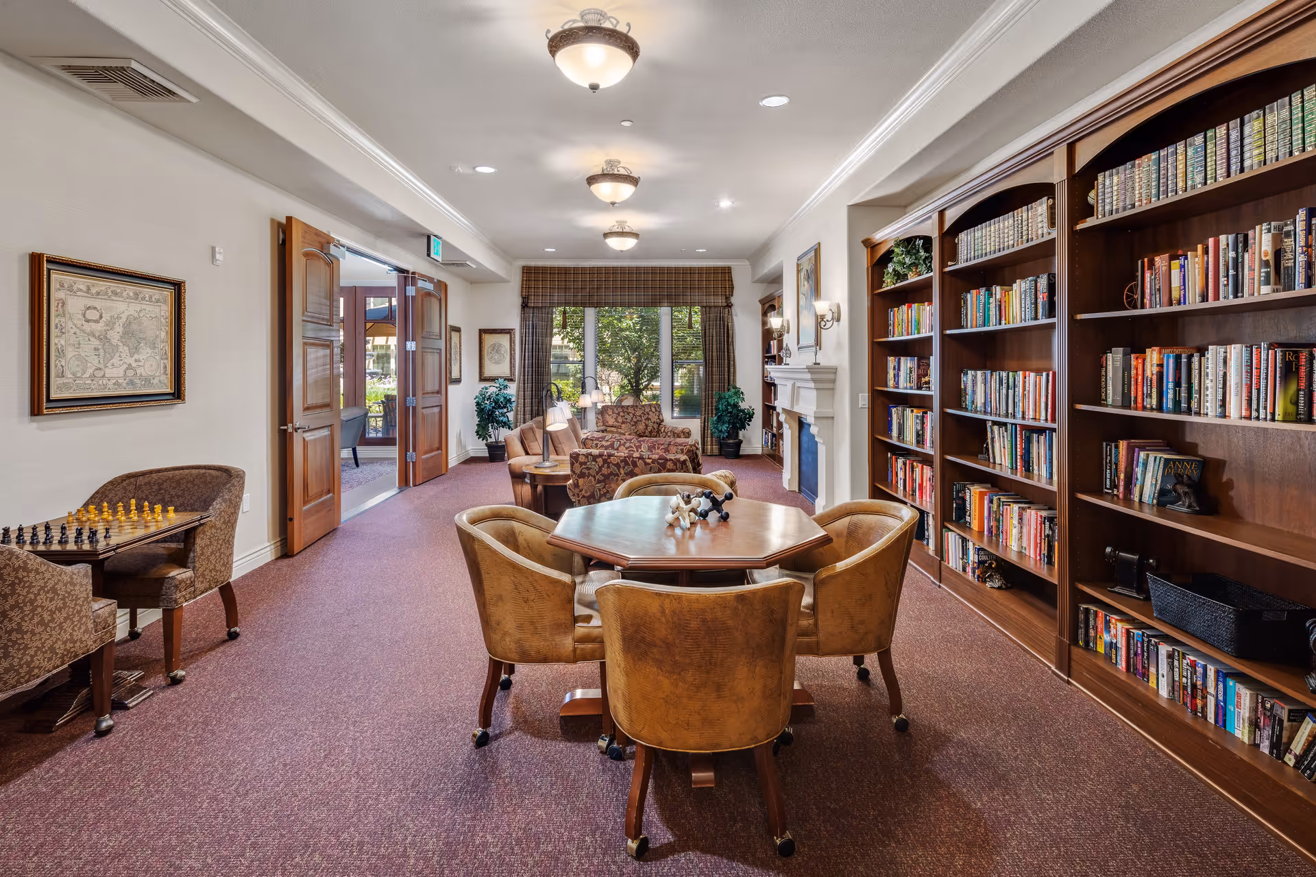 A cozy library and lounge area in a senior living facility with wooden bookshelves filled with books on the right, a hexagonal table with four chairs in the center, and a chess table with two chairs on the left. The room has carpeted floors, a fireplace, and large windows with curtains letting in natural light. Double wooden doors lead to an outdoor area.
